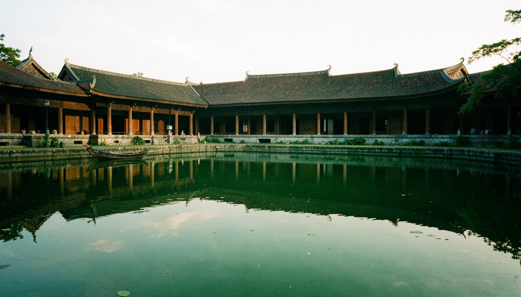 A wide view of the temple ordination hall seen across a still reflecting pond, the boat-shaped wall silhouette mirrored in the water under a soft morning sky.