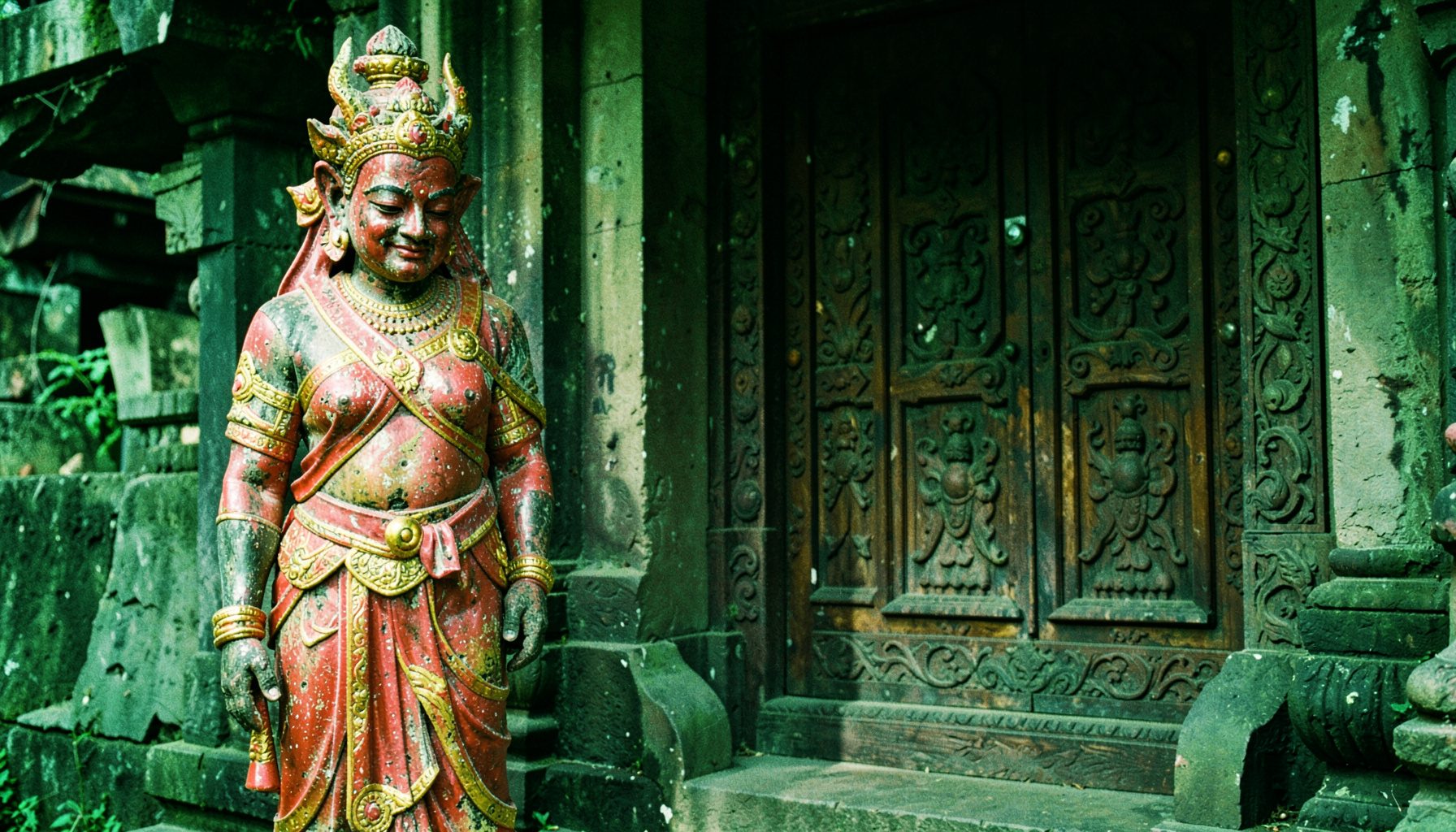 A weathered guardian statue at the temple entrance, painted in faded red and gold, standing watch beside a carved wooden door.
