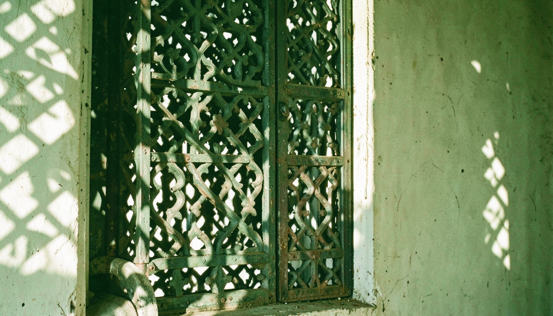 Detail of a window panel with intricate carved latticework, casting geometric shadows across a sun-bleached wall.