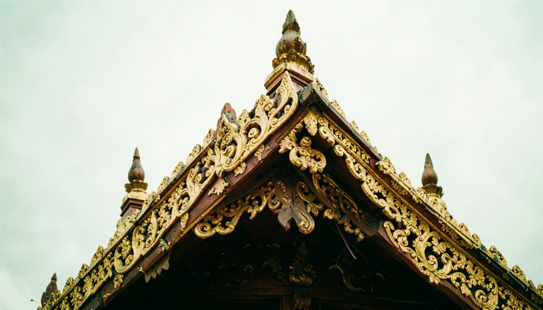 Close-up of an ornate temple gable with gold leaf detailing and carved wooden finials against a pale overcast sky, warm browns and muted golds.