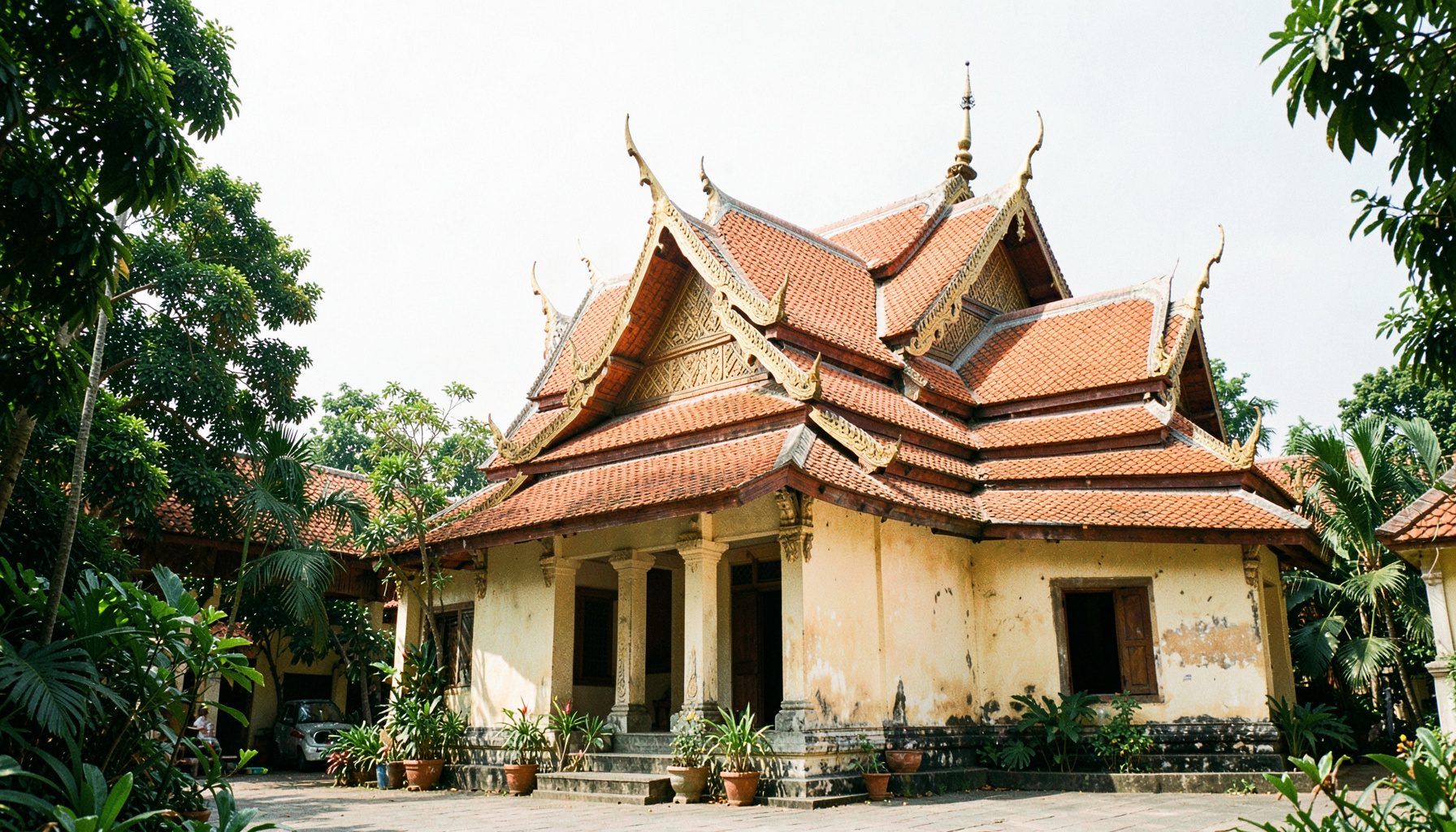 A sunlit temple courtyard with a traditional Thai ordination hall featuring layered orange-tiled roofs, ornate gable finials, and a weathered stucco facade in warm cream tones, framed by tropical foliage under a hazy sky.