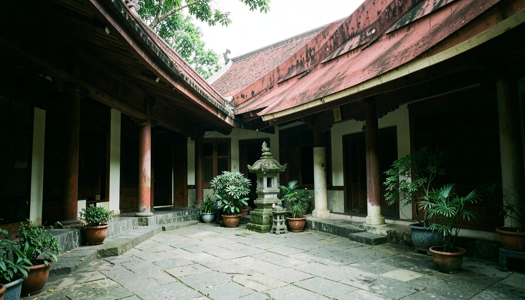 A quiet temple courtyard with stone paving, a small shrine, and potted plants, framed by the sloping roof eaves in deep terracotta and cream.